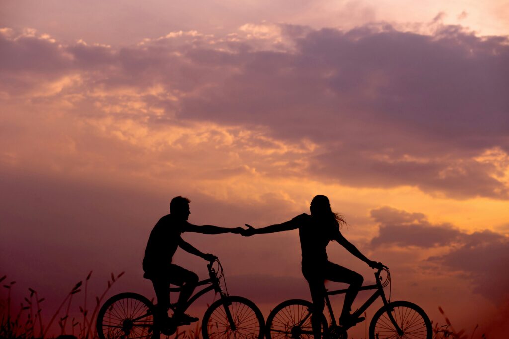 Two people ride bicycles at sunset, extending their arms to hold hands, with a cloudy sky and tall grass in the background.