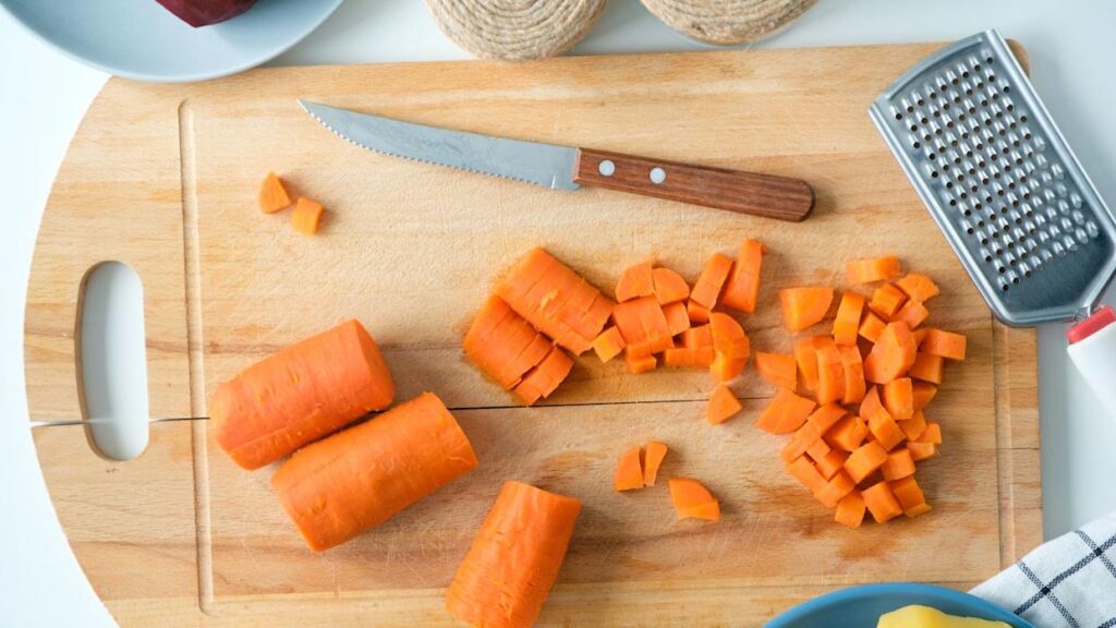 A wooden cutting board with chopped and whole carrots, a serrated knife, and a metal grater on a white surface.