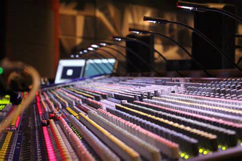 A close-up view of a professional audio mixing console with various colored buttons, sliders, and small reading lights in a recording studio.