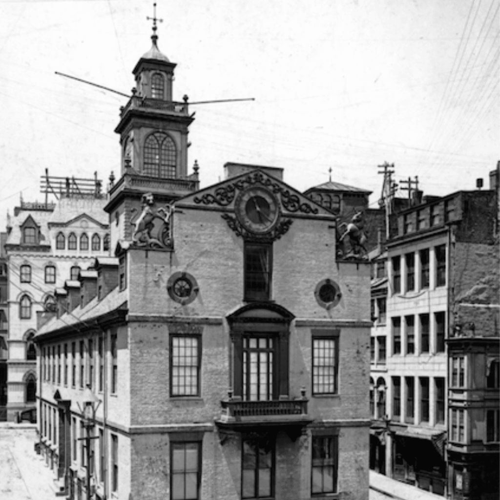 Black-and-white photo of an old multi-story building with a clock and statues on the facade, surrounded by other historic city buildings.