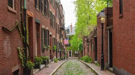 A narrow cobblestone street lined with brick buildings, green plants, and an American flag hanging in the distance.