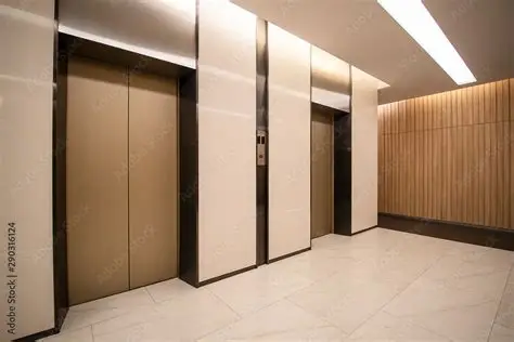 Two closed elevator doors in a modern, well-lit building lobby with light-colored walls, tiled floor, and wood panel accent.