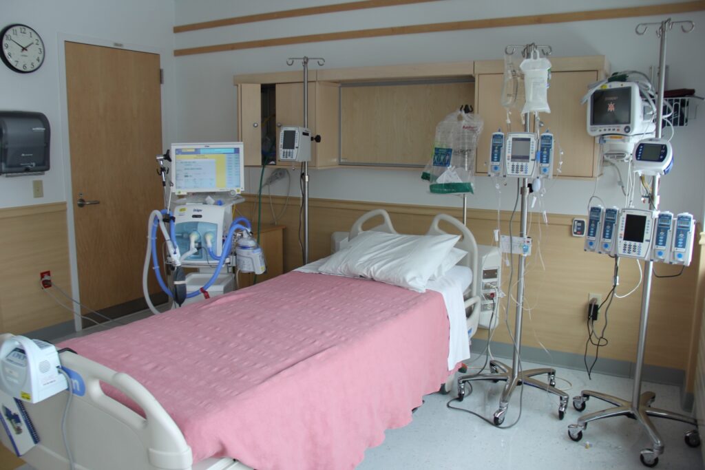 A hospital room with a bed covered by a pink blanket, surrounded by medical monitors, IV poles, and equipment. The room is empty with a clock, cabinets, and a closed door visible.