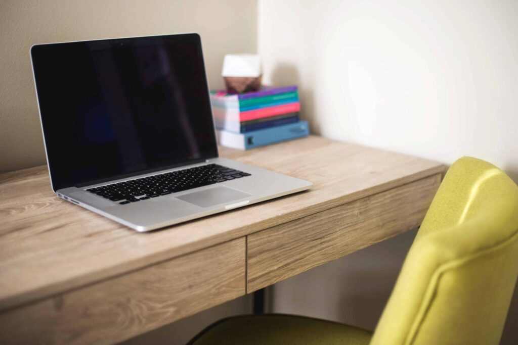 A closed laptop sits on a wooden desk next to a stack of colorful books, with a lime green chair in front.