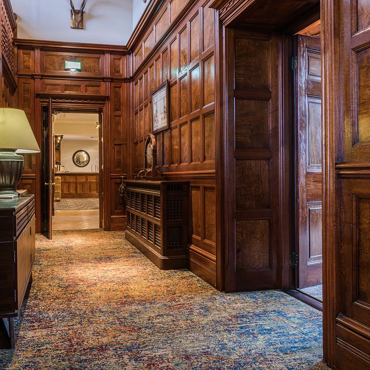 A wooden-paneled hallway with patterned carpet, a table lamp on a side table, and an open door leading to another well-lit room. A mounted animal head decorates the wall above.