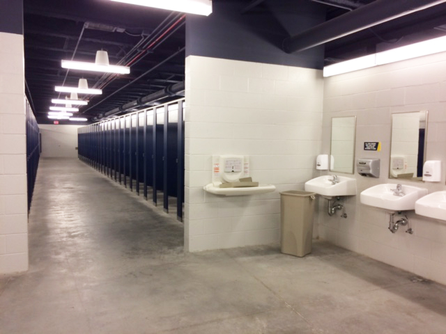 A large public restroom with multiple stalls along one wall, two sinks and mirrors, soap dispensers, and a trash can in the foreground.