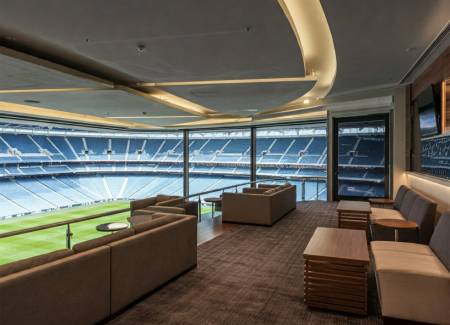 A modern stadium suite with sofas and tables overlooks an empty sports field and rows of blue stadium seats through large windows.
